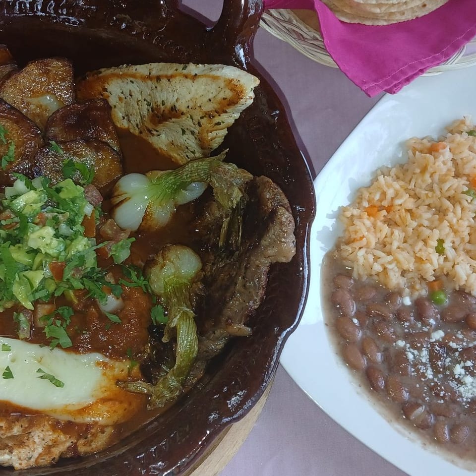 Mexican-style platter with grilled steak, vegetables, potatoes, melted cheese, rice, and refried beans on a table.